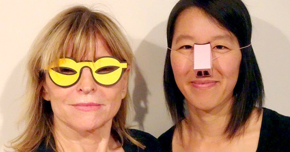 Eine deutsch aussehende, blonde Frau mit einer gelben Schlitzaugenbrille und eine asiatisch aussehende, dunkelhaarige Frau mit einer rosa Pappnase stehen nebeneinander und schauen freundlich in die Kamera.