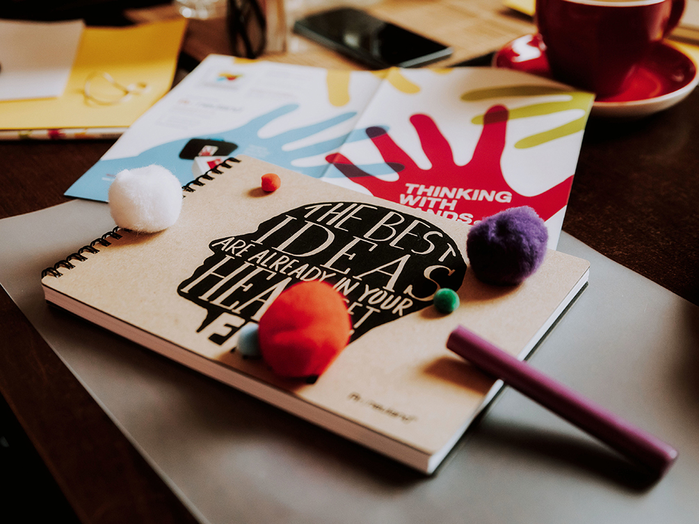 Ein Schreibblock mit einem Kopf und der Aufschrift „The best ideas are already in your head – set them free“, bunte Kreativmaterialien und ein Faltblatt mit dem farbigen Logo von „Thinking with Hands“ liegen wohl platziert auf der Arbeitsfläche.