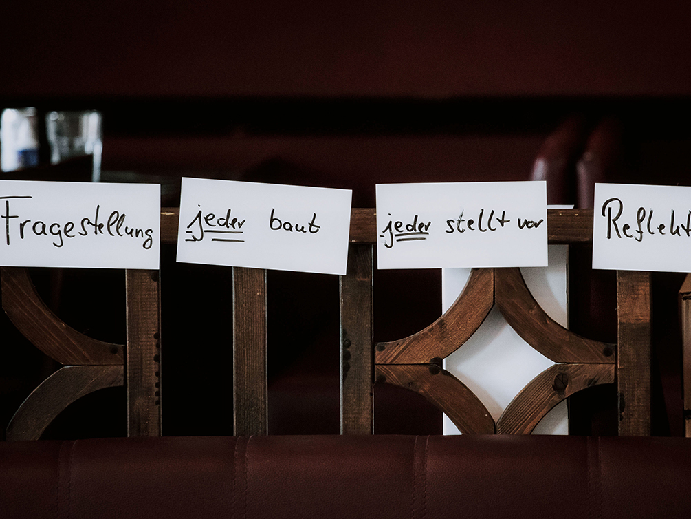 Vier Moderationszettel mit den Begriffen „Fragestellung“, „jeder baut“, „jeder stellt vor“ und „Reflektionen“ haften nebeneinander an einem Geländer.