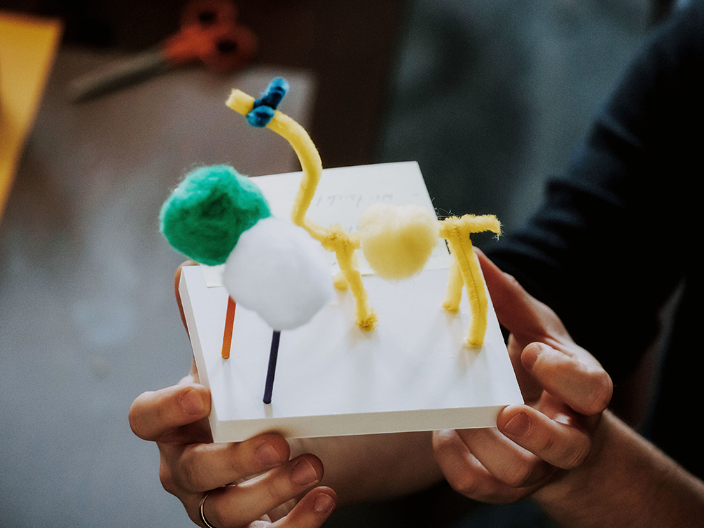 Zwei Hände halten ein Modell. Das Modell besteht aus einer Sandwich-Platte, gelben und blauen Pfeifenreiniger und zwei unterschiedlich farbige Stöckchen, worauf jeweils ein grünes und ein weißes Pompon aufgesetzt ist.