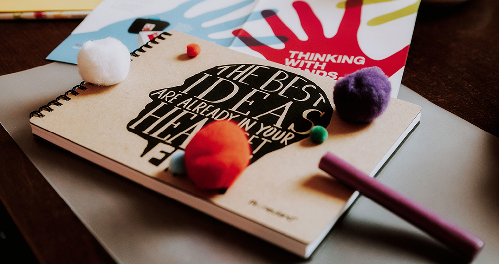 Ein Schreibblock mit einem Kopf und der Aufschrift „The best ideas are already in your head – set them free“, bunte Kreativmaterialien und ein Faltblatt mit dem farbigen Logo von „Thinking with Hands“ liegen wohl platziert auf der Arbeitsfläche.