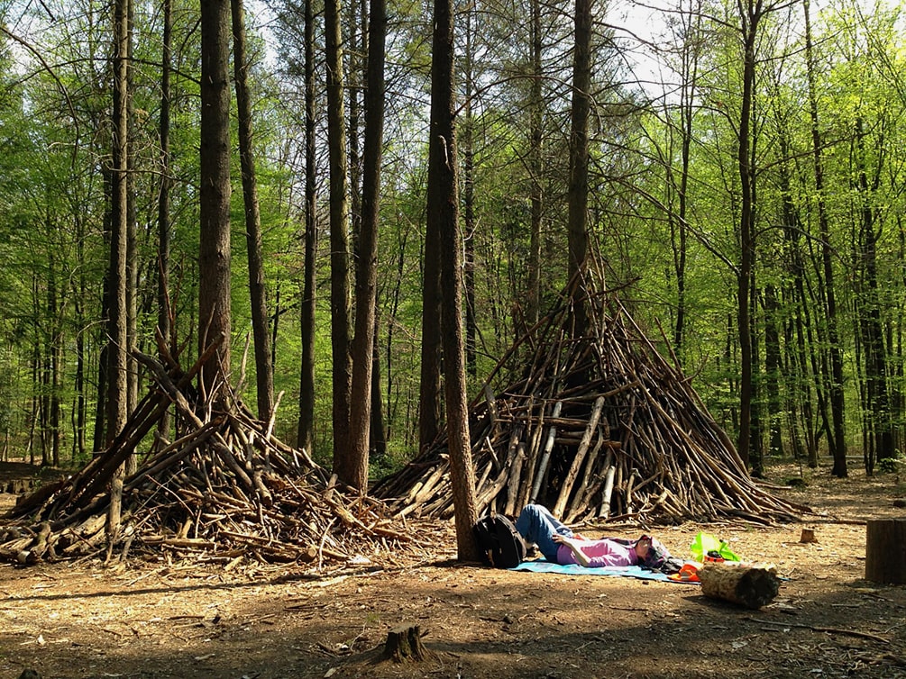 Auf eine Waldlichtung liegt eine Frau auf einer Picknickdecke und macht ein Nickerchen. Im Hintergrund befinden sich zwei aus Ästen gebaute Höhlen. Die Bäume weisen frisches, grünes Laubwerk auf.